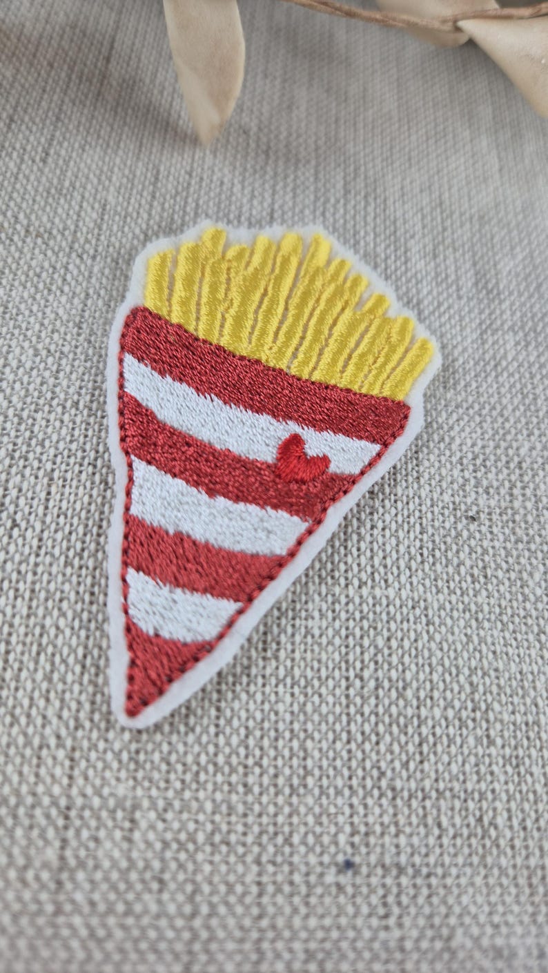 May include: Embroidered patch in the shape of a french fry container. The top is yellow, representing fries, and the bottom is red and white striped. A small red heart is embroidered on the front.