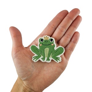 Puede incluir: Rana verde bordada con una corona dorada, sentada sobre un fondo blanco.