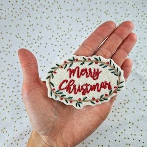 Könnte beinhalten: Ein weißer Filz-Weihnachtsanhänger mit einem grünen Kranz und roten Beeren, die den Text "Frohe Weihnachten" umgeben.