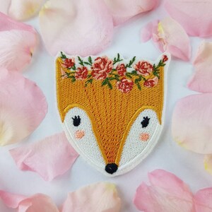 Può includere: Un toppa ricamata da stirare con un volpe con una corona di fiori. La volpe è arancione con dettagli bianchi e occhi neri. I fiori sono rosa e verdi.