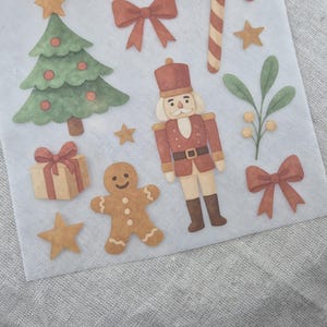 Puede incluir: Ilustraciones de acuarela con temática navideña sobre un fondo blanco. La imagen incluye un árbol de Navidad, una caja de regalo, un hombre de jengibre, un cascanueces, un bastón de caramelo, lazos y estrellas. Las ilustraciones son en tonos verdes, rojos, marrones y dorados.