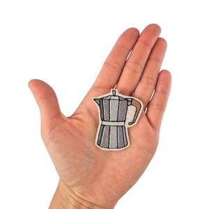 Może przedstawiać: Naszywka z haftowanej żelaznej kawiarki w kolorze szarym i brązowym. Naszywka znajduje się na dłoni.