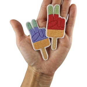 Puede incluir: Dos parches bordados para planchar con forma de polos con signos de paz. El polo de la izquierda es morado, naranja y verde. El polo de la derecha es rojo, naranja y verde.