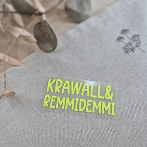 Könnte beinhalten: Ein transparenter Aufkleber mit dem Text "KRAWALL & REMMIDEEMI" in leuchtend gelben Buchstaben.