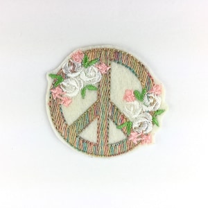 Könnte beinhalten: Gestickter Peace-Zeichen-Aufnäher mit floralem Design. Das Peace-Zeichen ist mit einem mehrfarbigen Faden umrandet, mit cremefarbenem Hintergrund. Weiße und rosa Blumen mit grünen Blättern sind auf den Aufnäher gestickt.