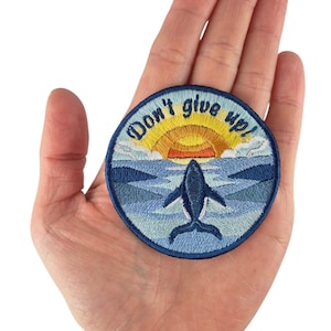 Könnte beinhalten: Gestickter Aufnäher mit blauem Rand, der einen Wal zeigt, der unter einem Sonnenuntergang aus dem Ozean springt. Der Aufnäher enthält den Text "Don't give up!" in einem kreisförmigen Design. Der Aufnäher wird in einer Hand gehalten.