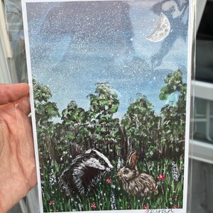May include: A rectangular art print featuring a painted scene of a badger and a rabbit in a grassy meadow with wildflowers. The background shows a dark forest under a starry night sky with a crescent moon. The artist signature Ryan is in the corner.