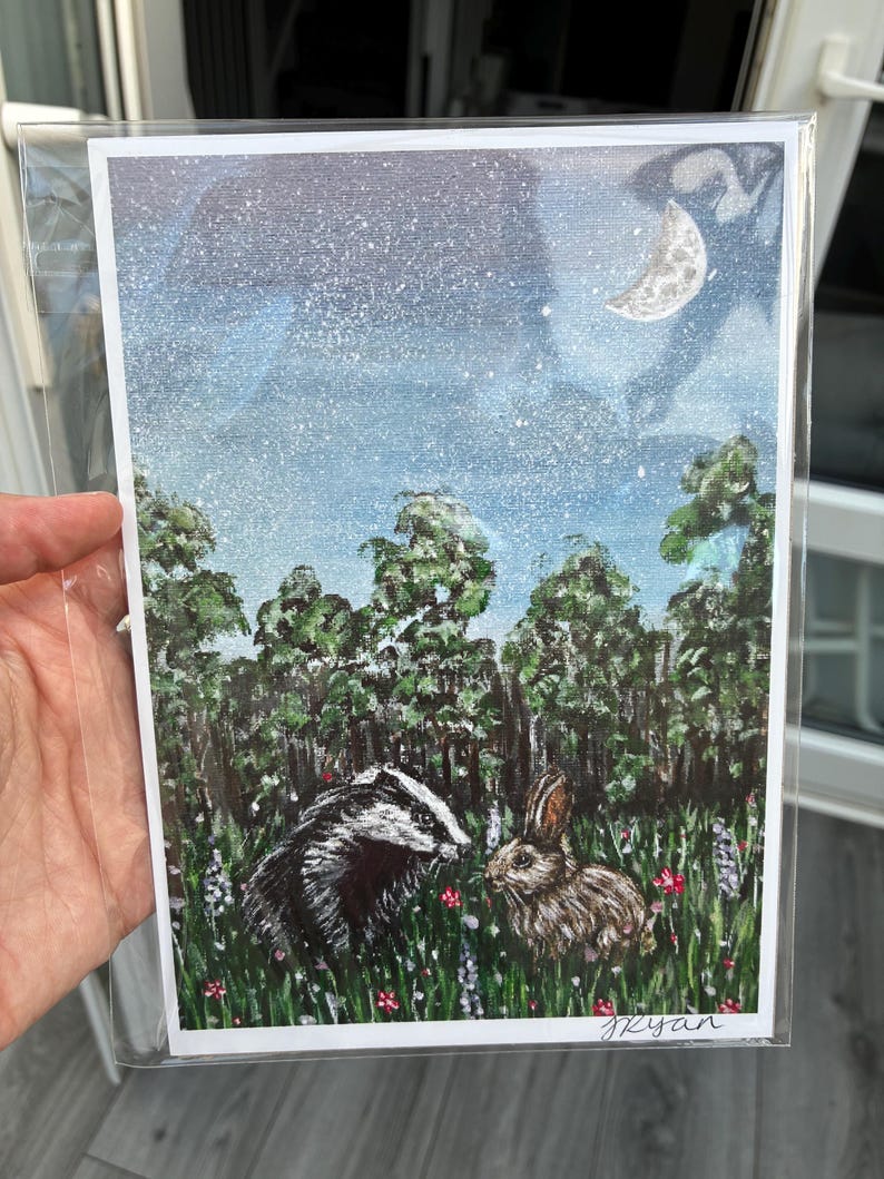 May include: A rectangular art print featuring a painted scene of a badger and a rabbit in a grassy meadow with wildflowers. The background shows a dark forest under a starry night sky with a crescent moon. The artist signature Ryan is in the corner.