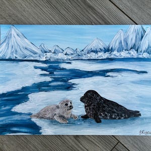 May include: Acrylic painting of two seals on an icy landscape. The painting features a light blue sky and white mountains in the background. One seal is gray, and the other is black. The painting is signed "J Ryan" in the bottom right corner.
