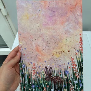May include: A rectangular canvas painting featuring a dark brown rabbit standing in a field of green grass and colorful wildflowers. The background is a soft, blended sky in shades of peach, pink, and yellow with scattered dark paint speckles.