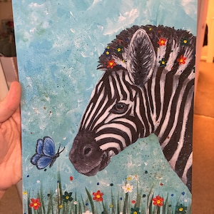 May include: Acrylic painting of a zebra's head adorned with flowers, set against a light blue background. A blue butterfly hovers near the zebra's face, with colorful flowers and grass in the foreground. The artwork is on a rectangular canvas.