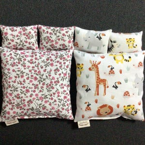 Könnte beinhalten: Eine Sammlung dekorativer Kissen. Einige Kissen zeigen ein Blumenmuster mit rosa Blüten und grünen Blättern auf weißem Grund. Andere Kissen haben Tierillustrationen, darunter eine Giraffe, ein Löwe und ein Elefant.