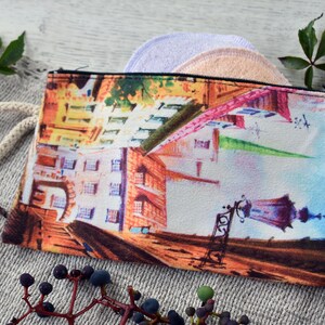 May include: A colorful zippered pouch with a watercolor cityscape design. The pouch has a white drawstring and a brown bead. The text "Latvia 100" is printed on the side of the pouch.