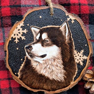 Può includere: Un ornamento in legno con un'immagine dettagliata di un cane Alaskan Malamute marrone e bianco bruciato a legno con fiocchi di neve sullo sfondo.