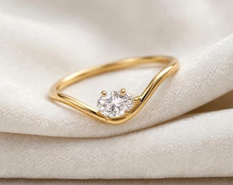 May include: A gold ring featuring a single, clear, oval-cut gemstone set in a prong setting. The band has a distinct wave or V-shaped curve, creating a delicate and asymmetrical silhouette that highlights the central stone's faceted surface.