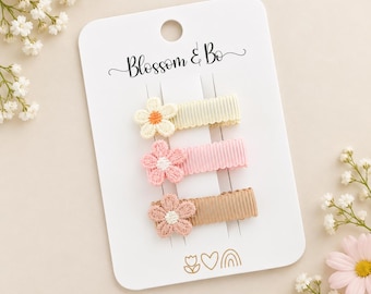 May include: Three hair clips on a white display card labeled Blossom & Bo. Each clip features a ribbed fabric band in cream, light pink, or tan, topped with a small embroidered flower in a matching or contrasting color. The card includes icons.