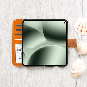 May include: A black smartphone in a case with a green and white abstract design, next to an orange wallet with card slots. A brown leather strap secures the phone. Seashells are scattered around the phone and wallet on a white wooden surface.