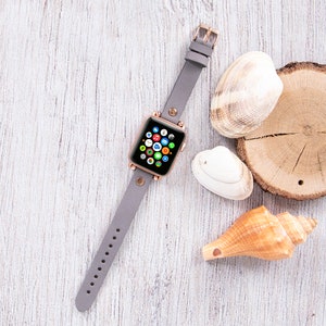 Könnte beinhalten: Ein graues Lederarmband für eine Apple Watch mit einem roségoldenen Zifferblatt und verschiedenen Apps, die auf dem Bildschirm angezeigt werden. Die Uhr liegt auf einer weißen Holzoberfläche mit Muscheln.