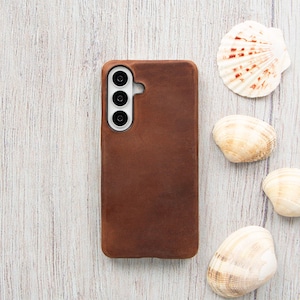 May include: A brown leather phone case with a triple-lens camera cutout. The case is rectangular and has a textured appearance. Several seashells are arranged around the phone case on a light-colored wooden surface.