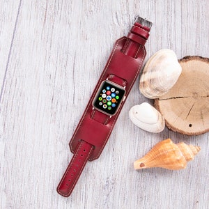 Könnte beinhalten: Ein rotes Lederarmband mit einem goldenen Apple Watch-Zifferblatt. Die Uhr liegt auf einer weißen Holzoberfläche mit Muscheln.