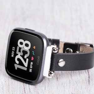 May include: A black leather band with silver accents for a Fitbit smartwatch. The watch face displays the time, date, and heart rate.