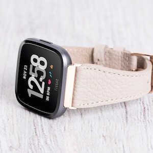 May include: A beige leather band with a rose gold buckle for a Fitbit Versa smartwatch. The watch face displays the time, date, and heart rate.