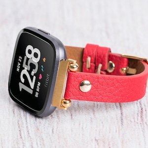 May include: A black Fitbit smartwatch with a red leather strap and gold accents. The watch face displays the time, date, and heart rate. The text on the watch face reads "NOV 23 12:58 96 BPM Fitbit".