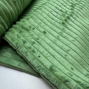 Könnte beinhalten: Ein salbeigrüner Cordstoff mit einer ausgeprägten vertikalen Rippenstruktur. Das Material weist einen weichen, flauschigen Flor mit durchgehenden parallelen Linien auf der gesamten Oberfläche auf und zeigt eine gefaltete Kante mit sichtbarer Dicke.