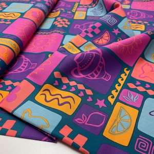 Op de afbeelding: Een levendige stof met een herhalend patroon van kleurrijke vierkanten. De vierkanten bevatten verschillende ontwerpen, waaronder zonnen, citroenen en schelpen, in tinten roze, oranje en paars tegen een teal achtergrond. De stof is gevouwen en toont zijn textuur.