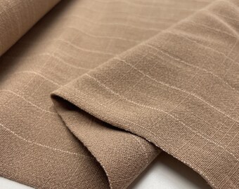 May include: A light brown fabric featuring a subtle grid pattern composed of thin, white, intersecting lines. The material has a visible woven texture and is draped in soft folds, highlighting the consistent check design across the surface.