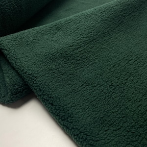 Könnte beinhalten: Ein dunkelwaldgrüner Stoff mit einer dicken, plüschigen und lockigen Sherpa-Fleece-Textur. Das Material ist umgeschlagen und zeigt den dichten, weichen und flauschigen Flor der synthetischen Textiloberfläche in einer tiefen, einheitlichen Farbe.