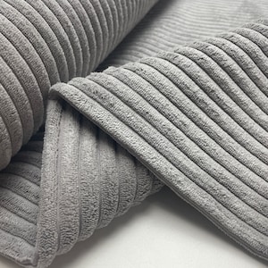 Op de afbeelding: Close-up van een grijze corduroy stof met een geribbelde textuur. De stof is dubbelgevouwen, waardoor de zachte, pluche textuur zichtbaar is.