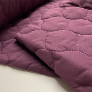 Peut inclure: Un tissu de couleur prune profonde présentant un motif matelassé avec des formes arrondies et imbriquées. Le matériau a une texture mate et douce avec des lignes de couture visibles créant un motif en grille festonné répétitif.