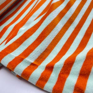 Könnte beinhalten: Ein weicher, plüschiger Stoff mit einem sich wiederholenden Muster aus kräftigen, leuchtend orangefarbenen Streifen, die sich mit blass hellminzgrünen Streifen abwechseln. Das Material hat eine flauschige, samtartige Textur und ist gefaltet.