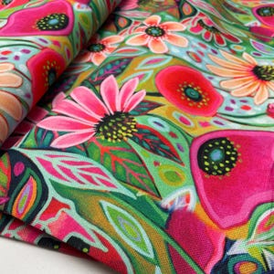 Op de afbeelding: Close-up van een levendige bloemenstof met grote roze, rode en oranje bloemen met groene bladeren tegen een lichtblauwe en groene achtergrond. Het ontwerp heeft een schilderkunstige stijl, met zichtbare penseelstreken en een gedurfd kleurenpalet.