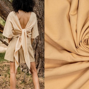 Op de afbeelding: Een beige tweedelige outfit met een top met een lage rug en een strikdetail, gecombineerd met een short. De outfit is gemaakt van een textuurstof. De afbeelding toont ook een close-up van de stof en de tekst "#milly".