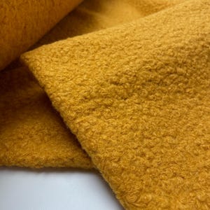 Op de afbeelding: Close-up van een getextureerde, goudgele stof. Het materiaal heeft een geluste, krullende textuur, met een zacht, pluche uiterlijk. De stof is opgevouwen en onthult de textuur en kleurvariaties.