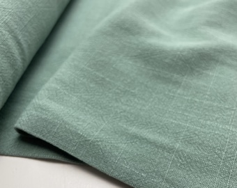 May include: Close-up of a folded piece of fabric in a muted, dusty green color. The fabric has a textured appearance with subtle woven details and a soft, matte finish. The folds create shadows and depth, highlighting the material's drape.