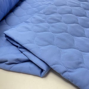 Könnte beinhalten: Ein gefalteter, hellblauer, gesteppter Stoff mit einem sich wiederholenden, wellenförmigen Stichmuster. Der Stoff hat eine einfarbige, hellblaue Farbe, und die Nähte sind in einem helleren Blauton gehalten. Der Stoff ist auf sich selbst gefaltet, wodurch die Textur und die Konstruktion des Materials sichtbar werden.