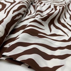 Op de afbeelding: Witte stof met een bruin zebra streepmotief. De stof is gevouwen en toont het herhalende patroon. De bruine strepen hebben een natuurlijke, organische vorm, die contrasteert met de witte achtergrond. Dit textiel is geschikt voor naai- en knutselprojecten.