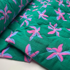 Op de afbeelding: Groene gewatteerde stof met een herhalend bloemenpatroon in roze en lichtblauw. De bloemen zijn gestileerd en gelijkmatig verdeeld. De stof is opgevouwen en toont de gewatteerde textuur.