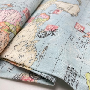 Könnte beinhalten: Hellblauer Stoff mit einem Vintage-Weltkarten-Design bedruckt. Die Karte zeigt Kontinente in Pastelltönen, mit roten Briefmarken-Illustrationen und schwarzem Text. Der Stoff ist gefaltet und zeigt den detaillierten Druck und die Textur.