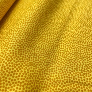 Op de afbeelding: Close-up van een gele stof met een herhalend patroon van kleine, gelijkmatig verdeelde, goudbruine stippen. De stof is licht gekreukt, waardoor schaduwen en kleurvariaties ontstaan. Het algehele effect is een gestructureerd, visueel interessant oppervlak.