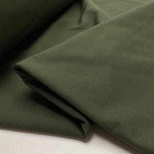 Op de afbeelding: Close-up van olijfgroene stof, gevouwen en gedrapeerd. De stof heeft een gladde textuur en een matte afwerking. De plooien creëren schaduwen en diepte, wat de drapering van de stof benadrukt.