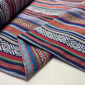 Può includere: Primo piano di un tessuto piegato con un vivace motivo a righe. Il design presenta bande alternate di blu navy, rosso, verde e bianco, con motivi geometrici e a forma di diamante. Il tessuto sembra essere tessuto.