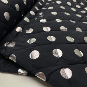 Op de afbeelding: Zwart gewatteerde stof met grote, zilverkleurige metallic stippen. De stof is opgevouwen, waardoor de textuur en het patroon zichtbaar zijn. Het quilten vormt een ruitpatroon. Geschikt voor naaiprojecten. Verkoop per meter.