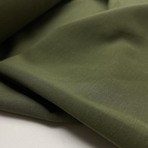 Op de afbeelding: Close-up van olijfgroene stof met zichtbare textuur en vouwen. De stof lijkt een gebreid of jersey materiaal te zijn, met een geribbelde rand zichtbaar in de linkerbenedenhoek. De kleur is een gedempt, aards groen.