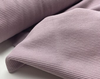May include: Close-up of a roll of mauve fabric. The material has a textured, waffle-like weave. The fabric is folded over itself, revealing the texture and colour variations. The material appears soft and pliable, suitable for clothing or home decor.