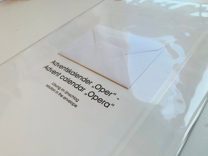 May include: A white envelope with the text "Adventskalender "Oper" - Advent calendar "Opera" L&ouml;sung im Umschlag solution in the envelope" printed on it.