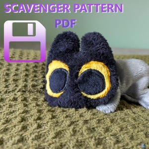May include: A plush toy in the shape of a creature with a black head, large yellow-rimmed eyes, and a gray body. The text "SCAVENGER PATTERN PDF" is displayed above the toy, along with a floppy disk icon.